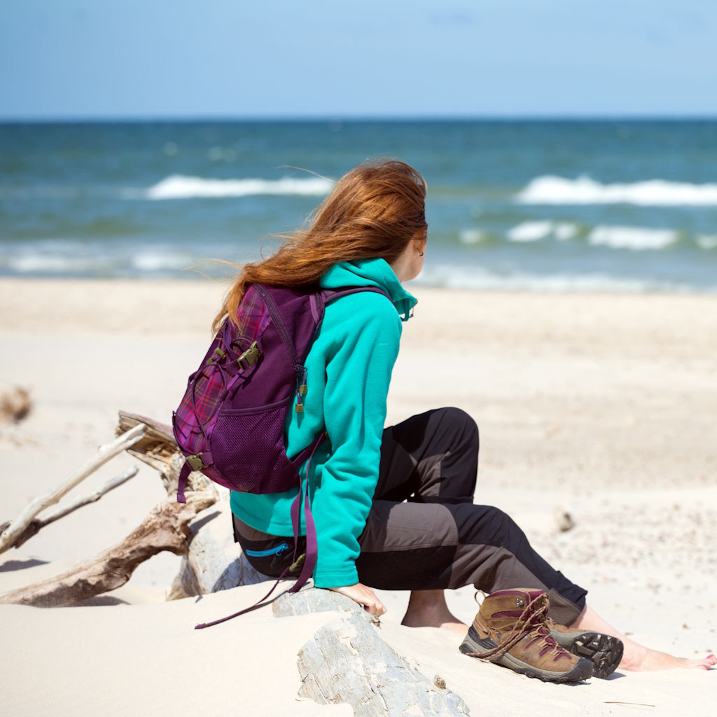 Frau sitzt auf Treibholz am Strand und hat ihre Wanderschuhe ausgezogen